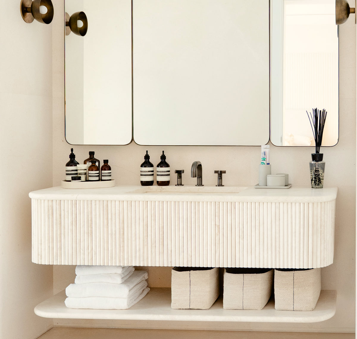 Modern bathroom vanity with floating sink, mirrors, neatly folded towels, and minimalist storage baskets.